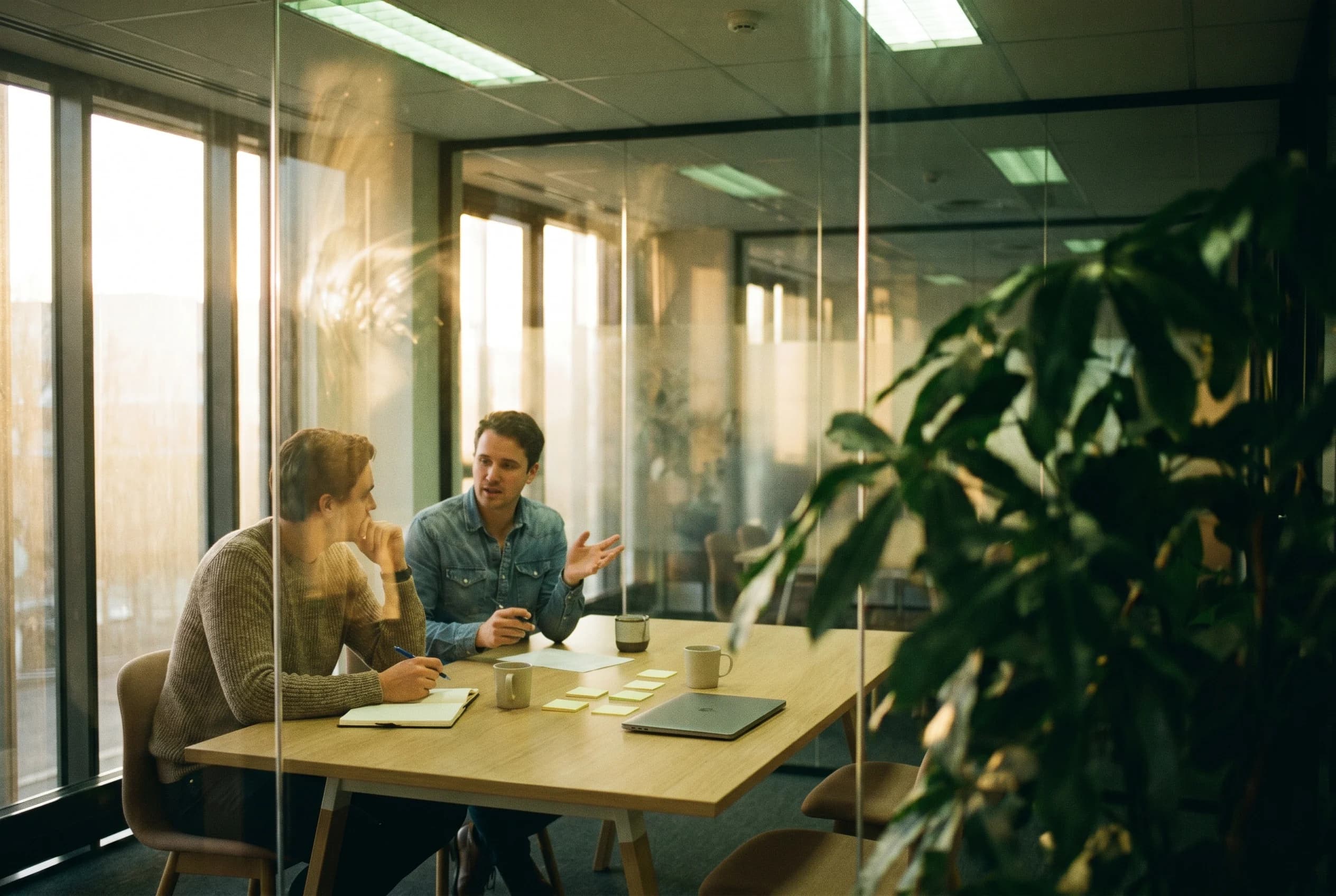 Two colleagues sit in a glass-walled meeting room during a calm discovery call practice session, with notebooks and warm window light.