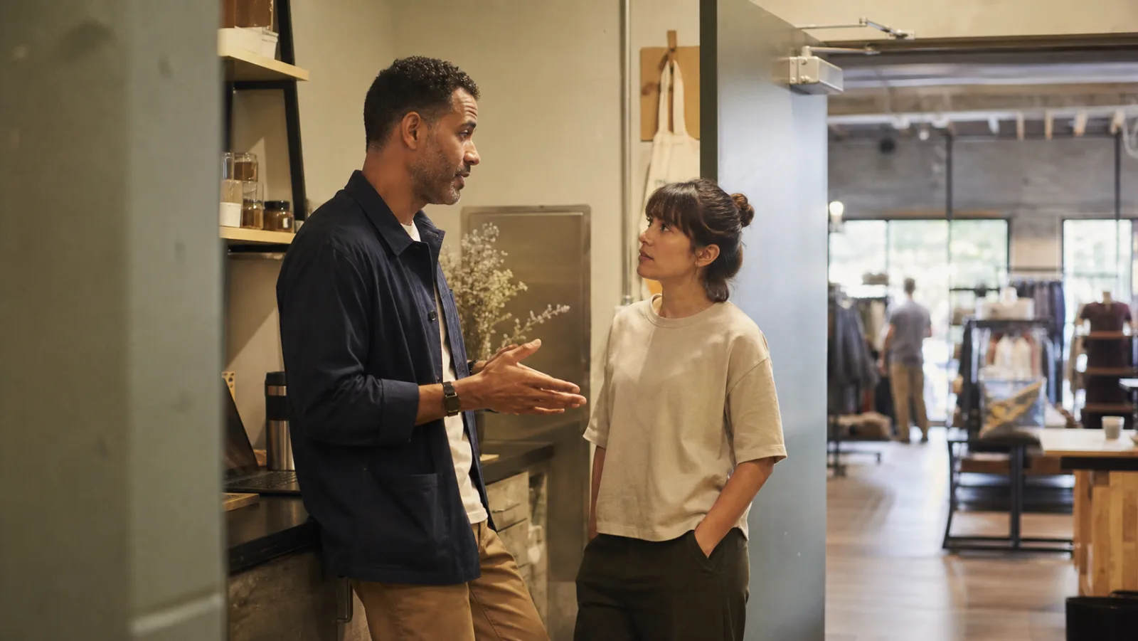 A retail manager and frontline associate have a calm coaching conversation in a warm back-of-house area beside the shop floor.